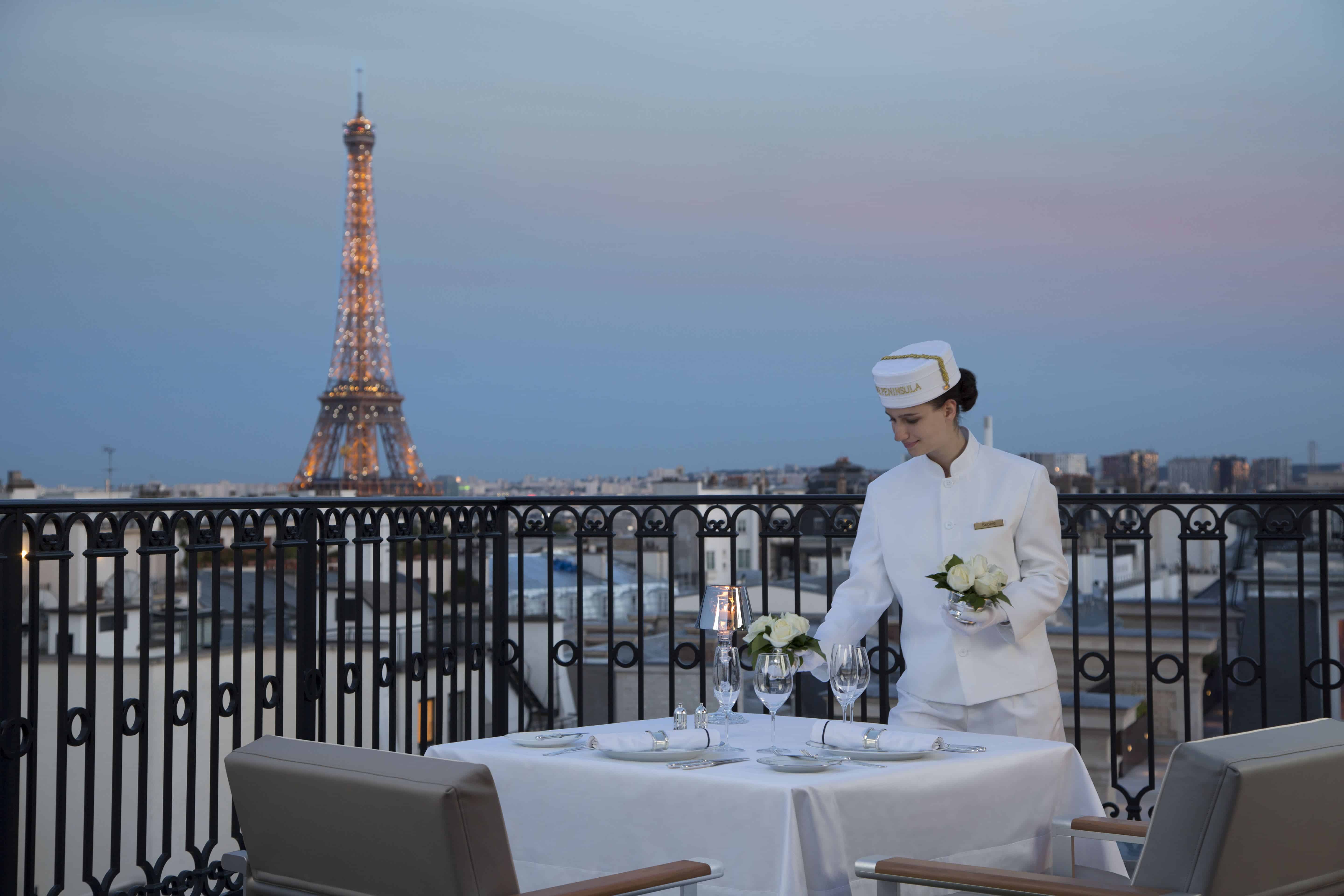 This photo is of a waiter in livery setting a beautiful table for a fancy dinner with an Eiffel Tower view. The Peninsula Paris is the top luxury hotel in Paris with Eiffel Tower view. 