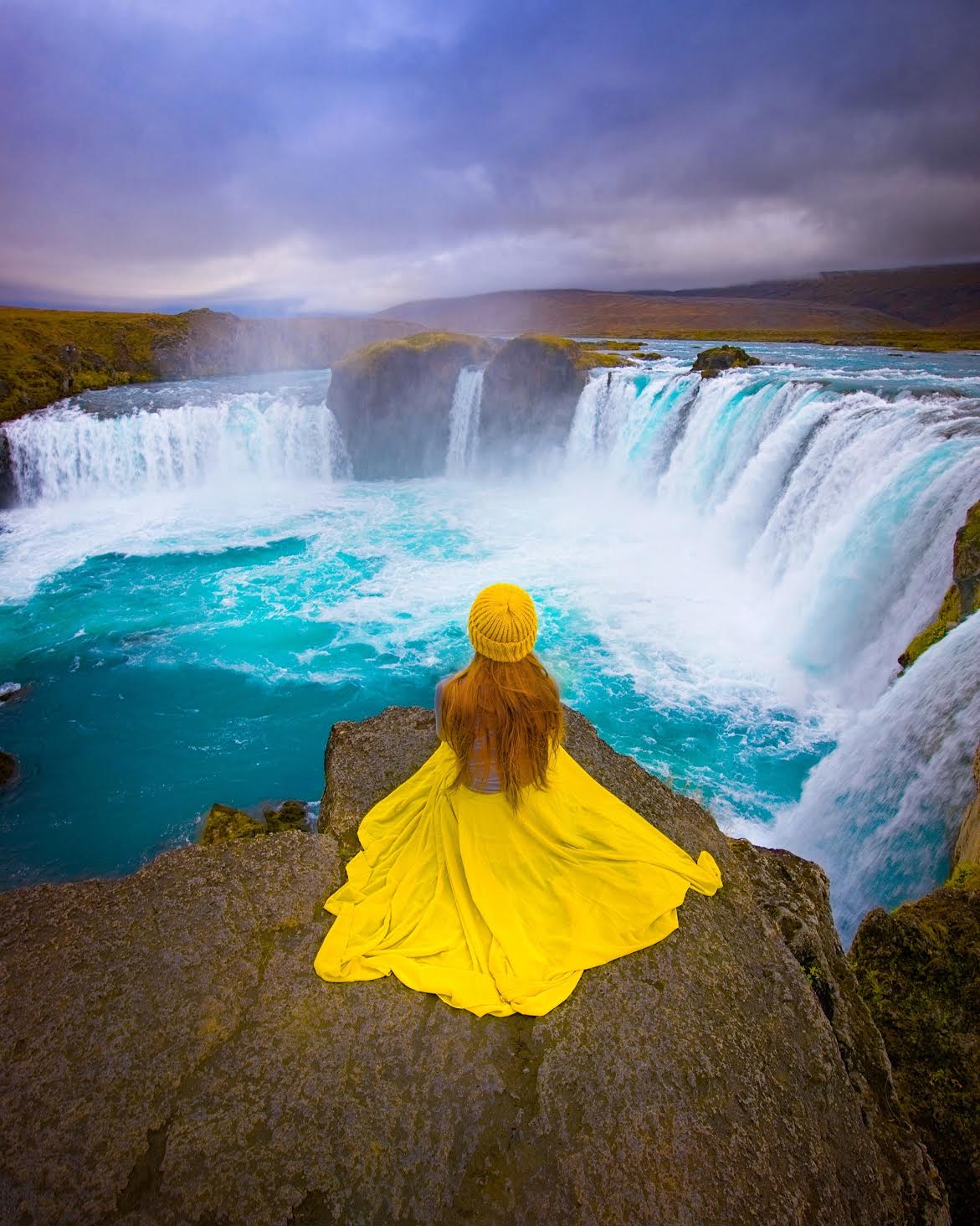 woman in a yellow skirt sitting with a waterfall in the background in iceland during march