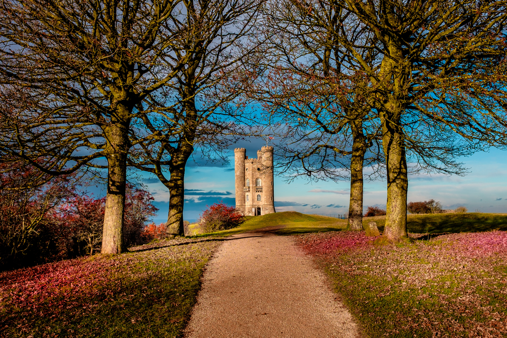 Broadway Tower with a path leading to and fallen leaves on the side of the path. A tree lined path leads to the tower. One of the places to vissit in the cotswolds. 