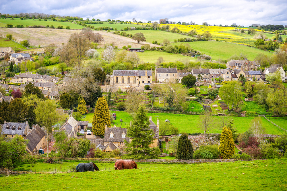 Picturesque View of Naunton Village, Cotswolds. There are horses in teh foreground and the village in the background.  