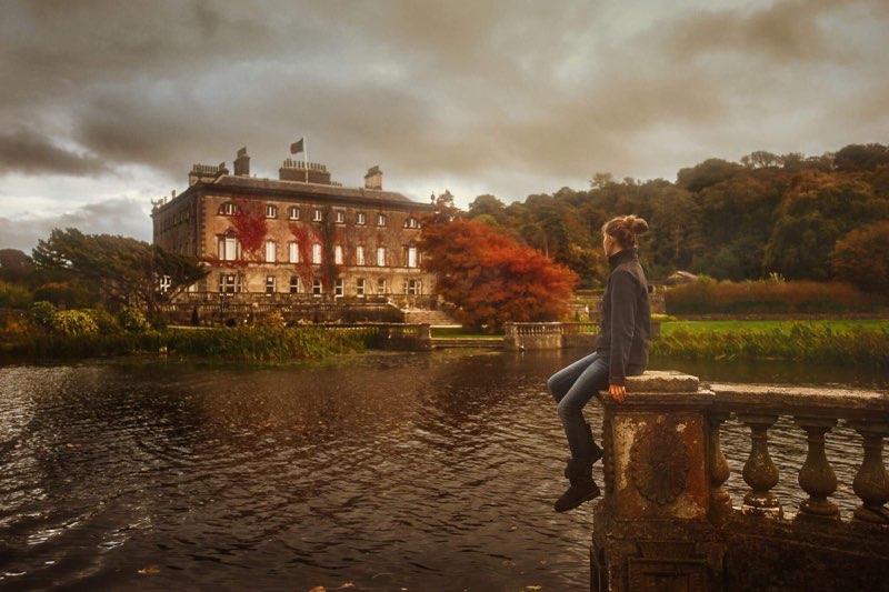 Young woman sitting on a stone fence next to a pond overlooking the Westport House on a fall overcast day.