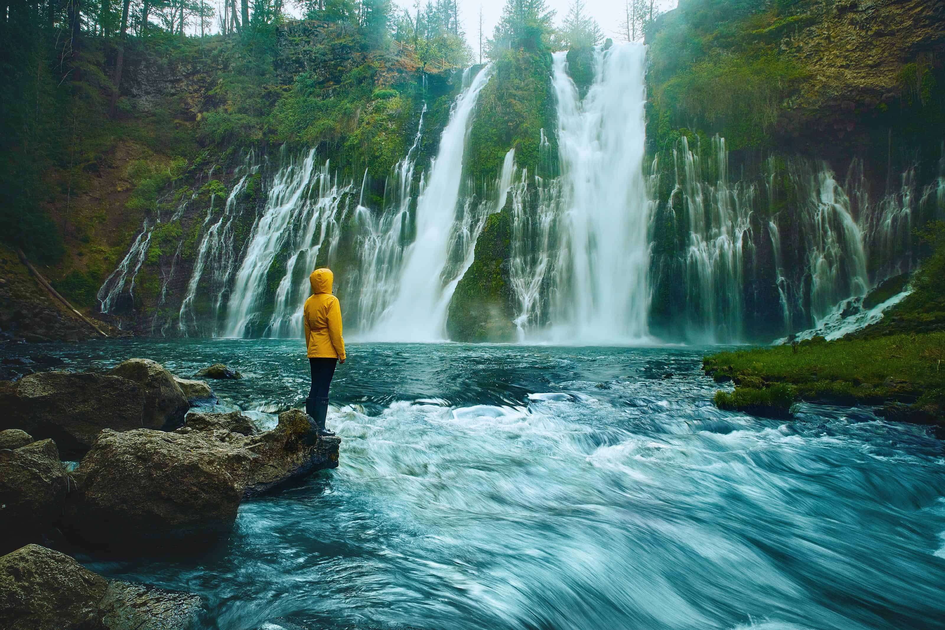 Figure in a yellow rain jacket standing on the edge of a river looking at Burney Falls with multiple streams on an overcast day.