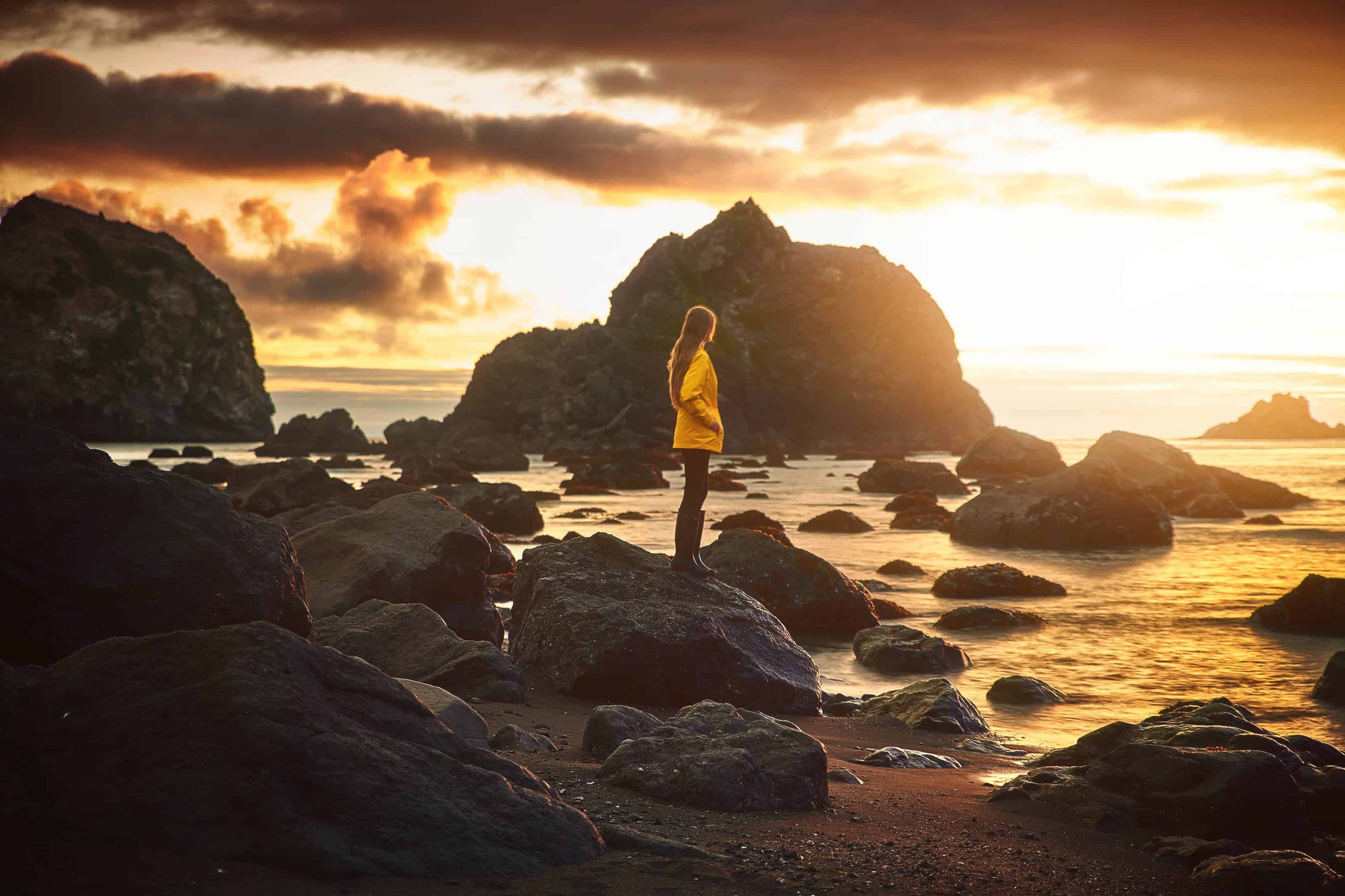 Woman in a yellow rain jacket standing on a rocky coast at sunset.