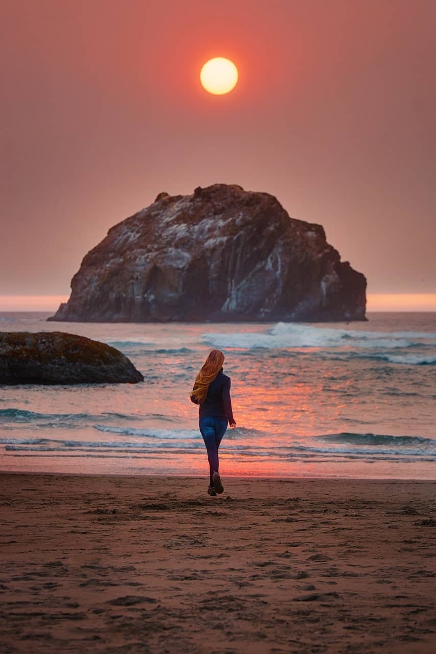 Woman with hair blowing in the wind walking at Face Rock in Bandon Oregon during pink sunset.