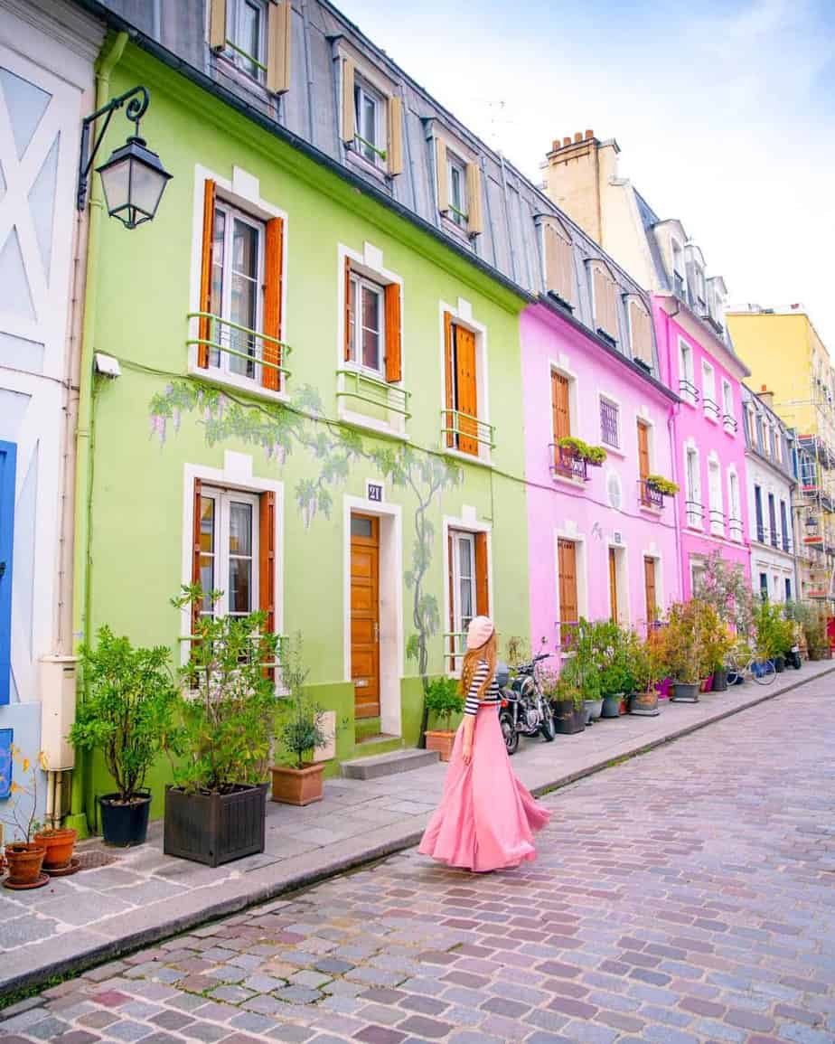 Woman in pink skirt and beret standing on Rue Cremiux with colorful houses and plants in Paris.