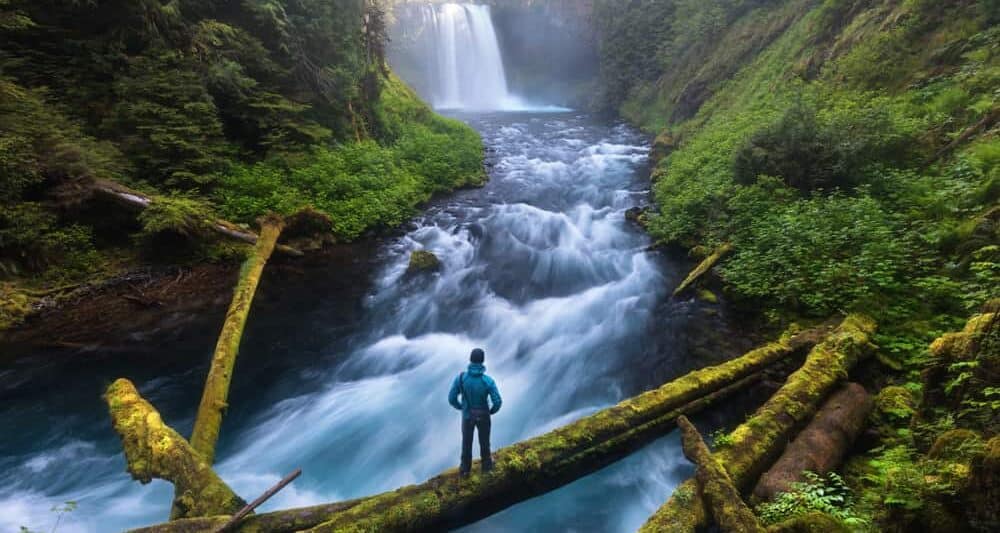 Man standing at the base of a rushing river with Koosah Falls in the background