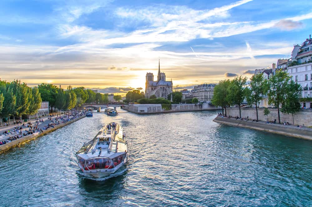 river boat sailing down the Seine river with Notre Dame in the background and with people along the shore of the river