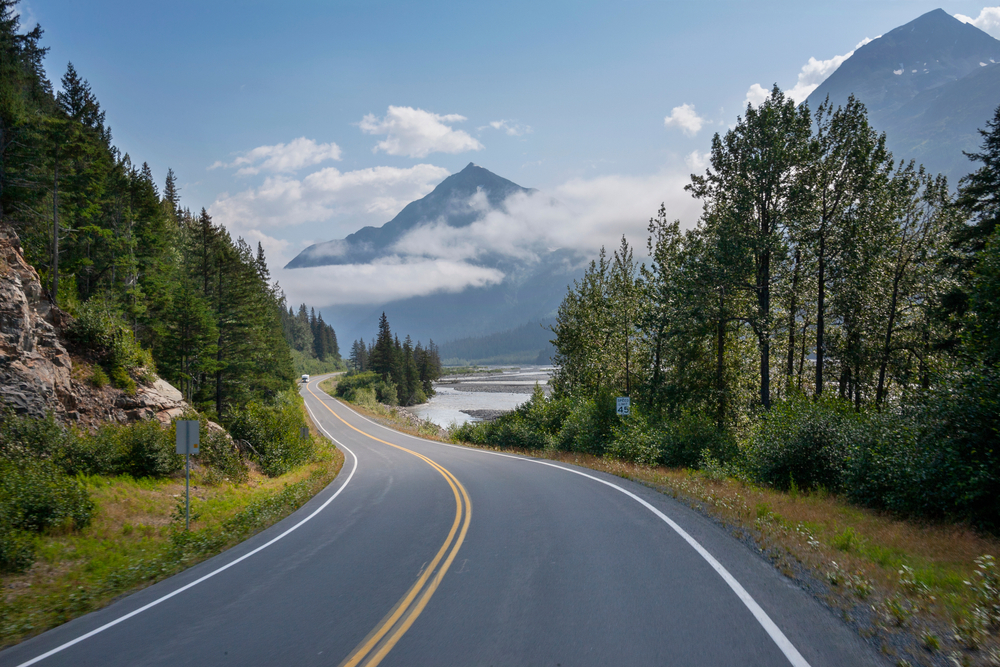 A winding road through trees heading toward mountains on an Alaska road trip.