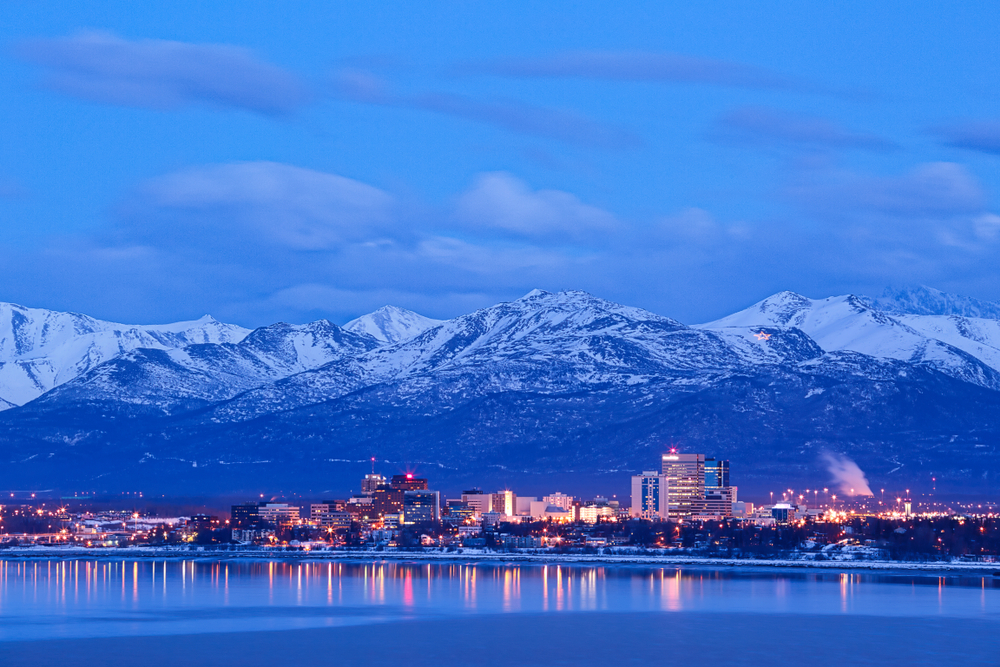 Blue hour over Anchorage with city lights reflecting in the water and snow-capped mountains in the distance.
