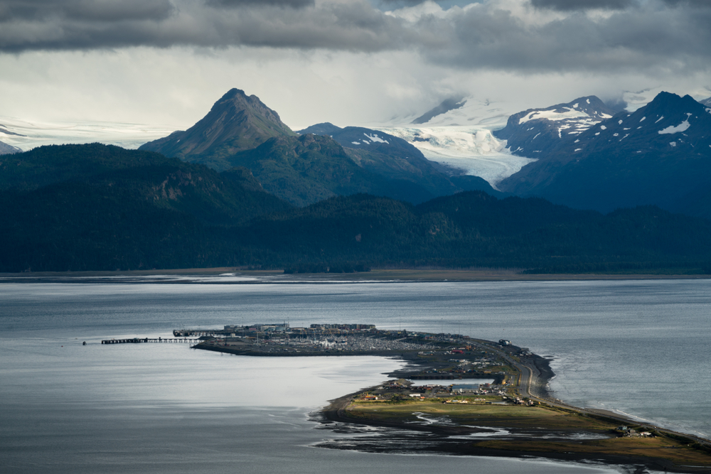 Aerial view of Homer on a strip of land in the water with mountains in the distance all under a cloudy sky.