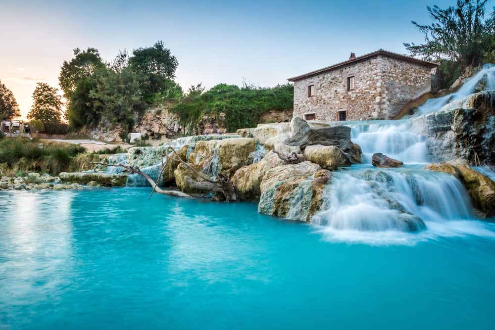 Pretty blue natural hot spring in Italy with waterfalls and stone building.