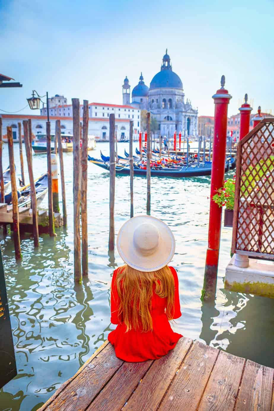 Woman in a red dress and sun hat sitting on the edge of a wooden dock looking across the water at the Basilica di Santa Maria della Salute, one of the best Instagrammable places in Venice.