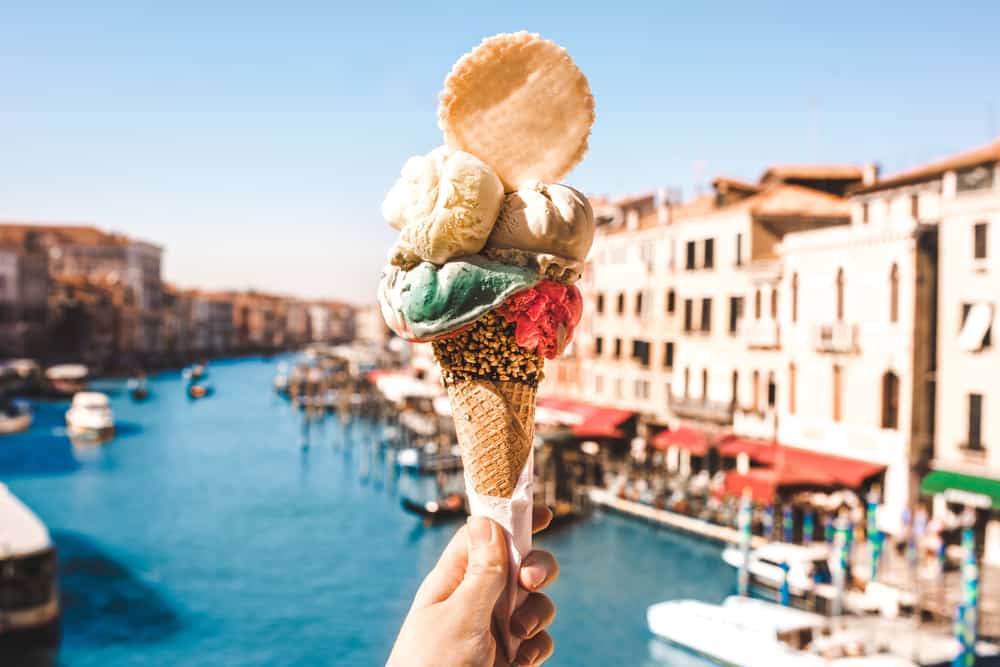 Hand holding a waffle cone piled with multiple colorful scoops of gelato over the Grand Canal in Venice.