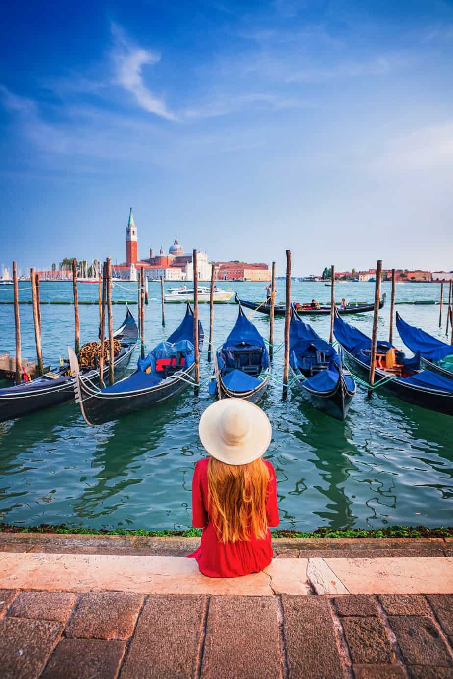 Woman in a red dress and sun hat sitting on the edge of the San Marco Waterfront with many blue docked gondolas and an island with a church in the distance.