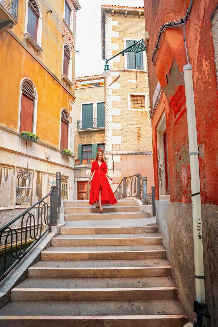 Woman in long, flowing red dress walking down the steps of a bridge among close buildings in Venice.