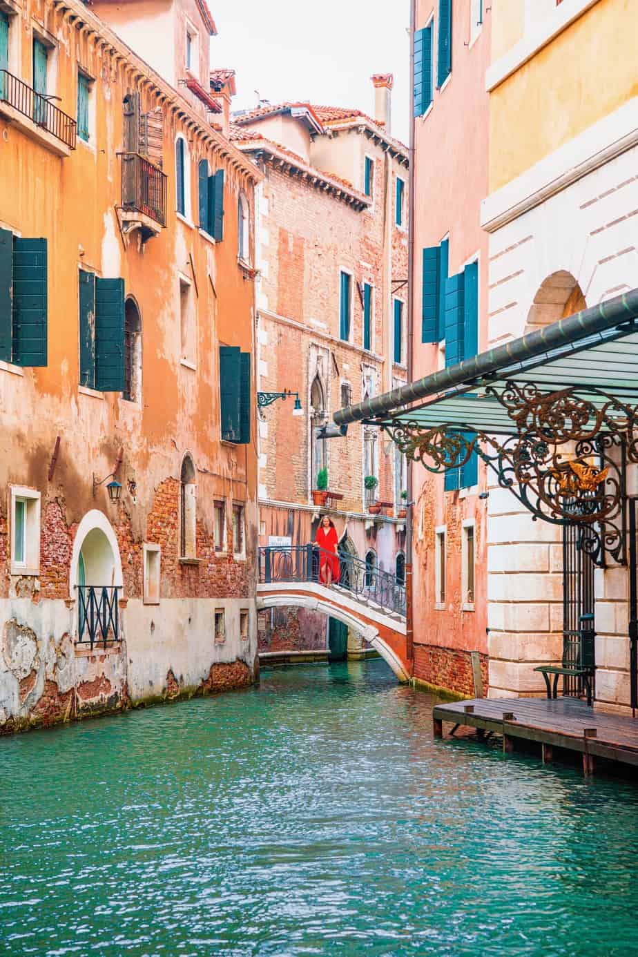 Shot of a woman in a red dress standing on a bridge between buildings over a canal in one of the most Instagrammable places in Venice.