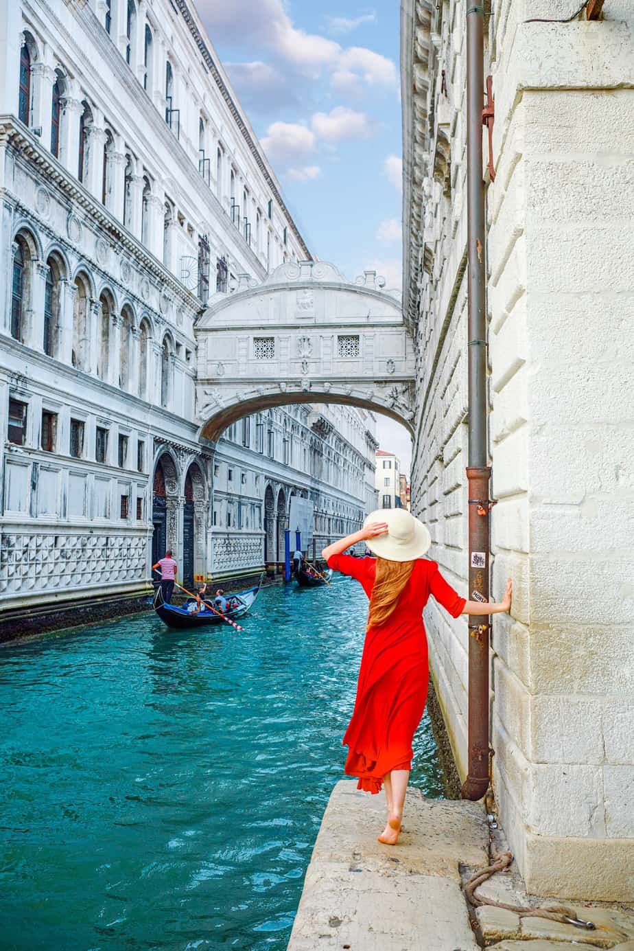 Woman in a red dress and sun hat stands under the white Bridge of Signs with gondolas on the canal.