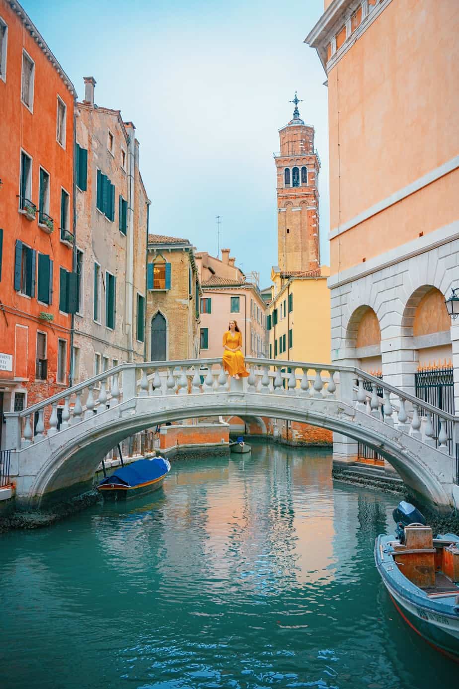 Woman in a yellow dress sitting on the rail of the Ponte Maria Callas over a canal with colorful buildings and a tower in the background.