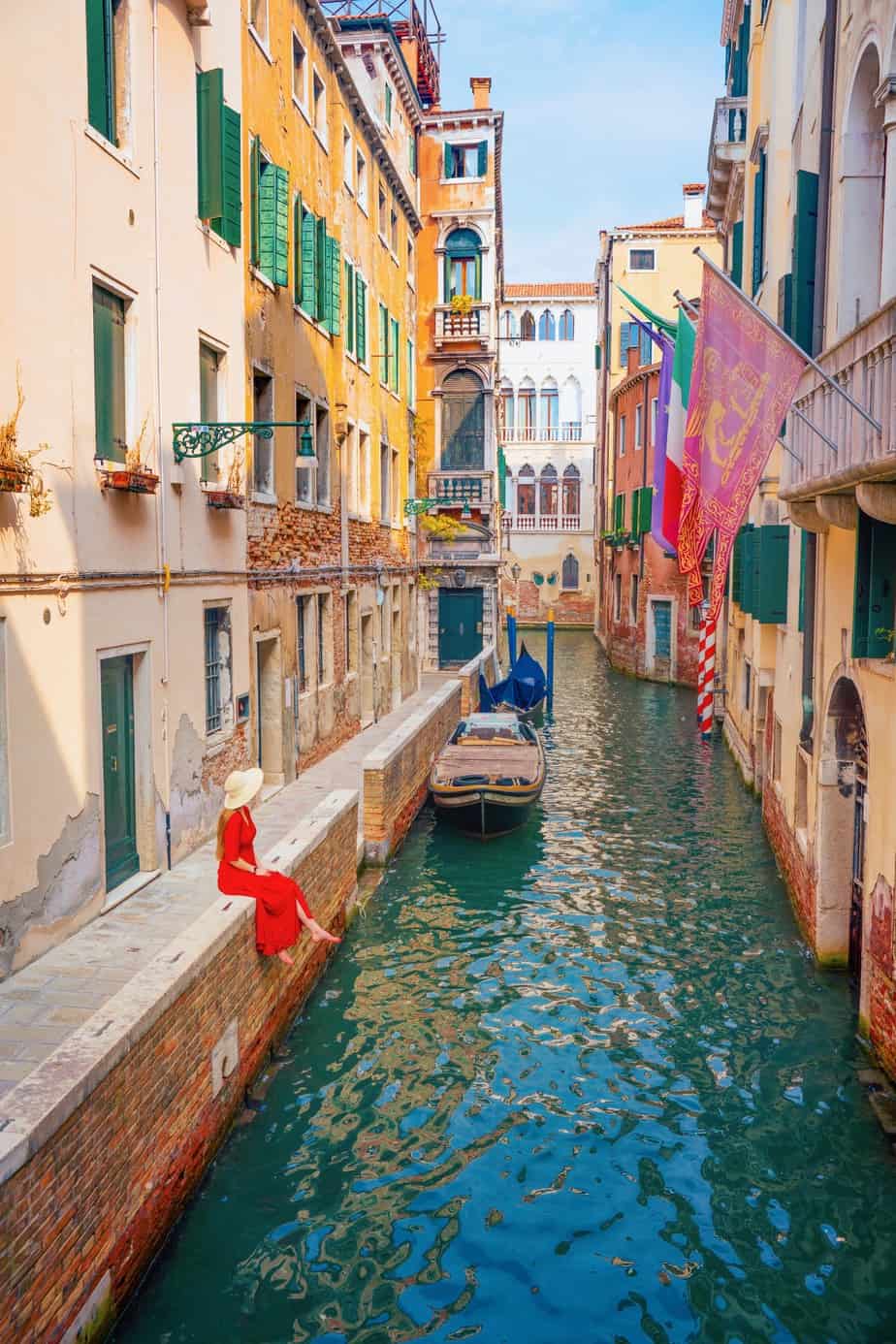 Looking down from Ponte de La Verona at a woman in a red dress and sun hat sitting on a brick wall on a canal with flags over the water.