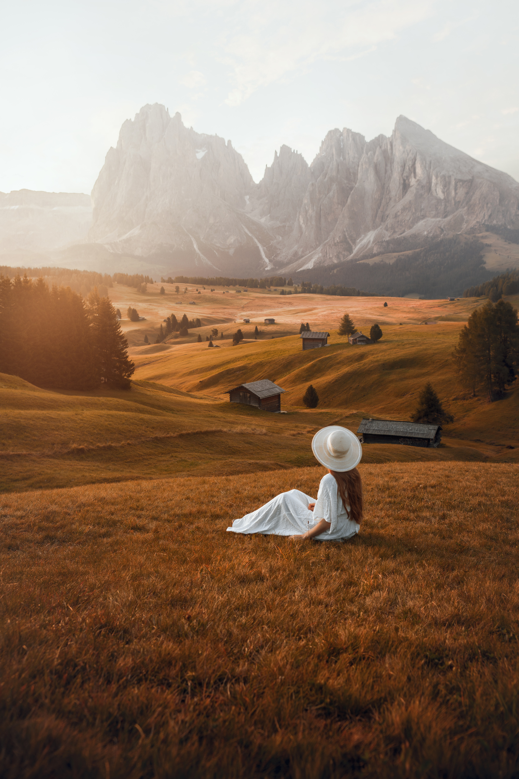 A women wearing a white dress and a hat sitting on tan plains in front of Dolomites mountain range.