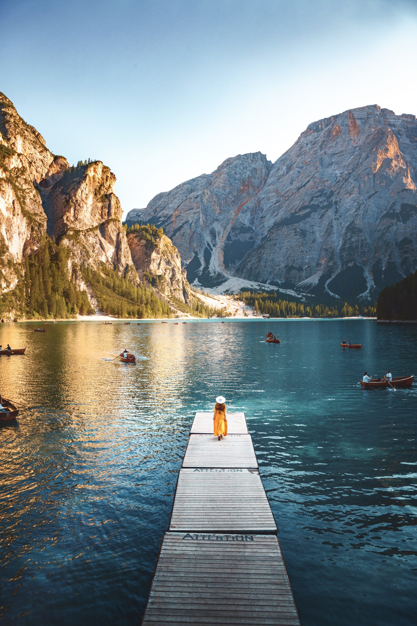 Aerial view of a woman standing on dock in the middle of turquoise lake with wooden boats and mountainous background.