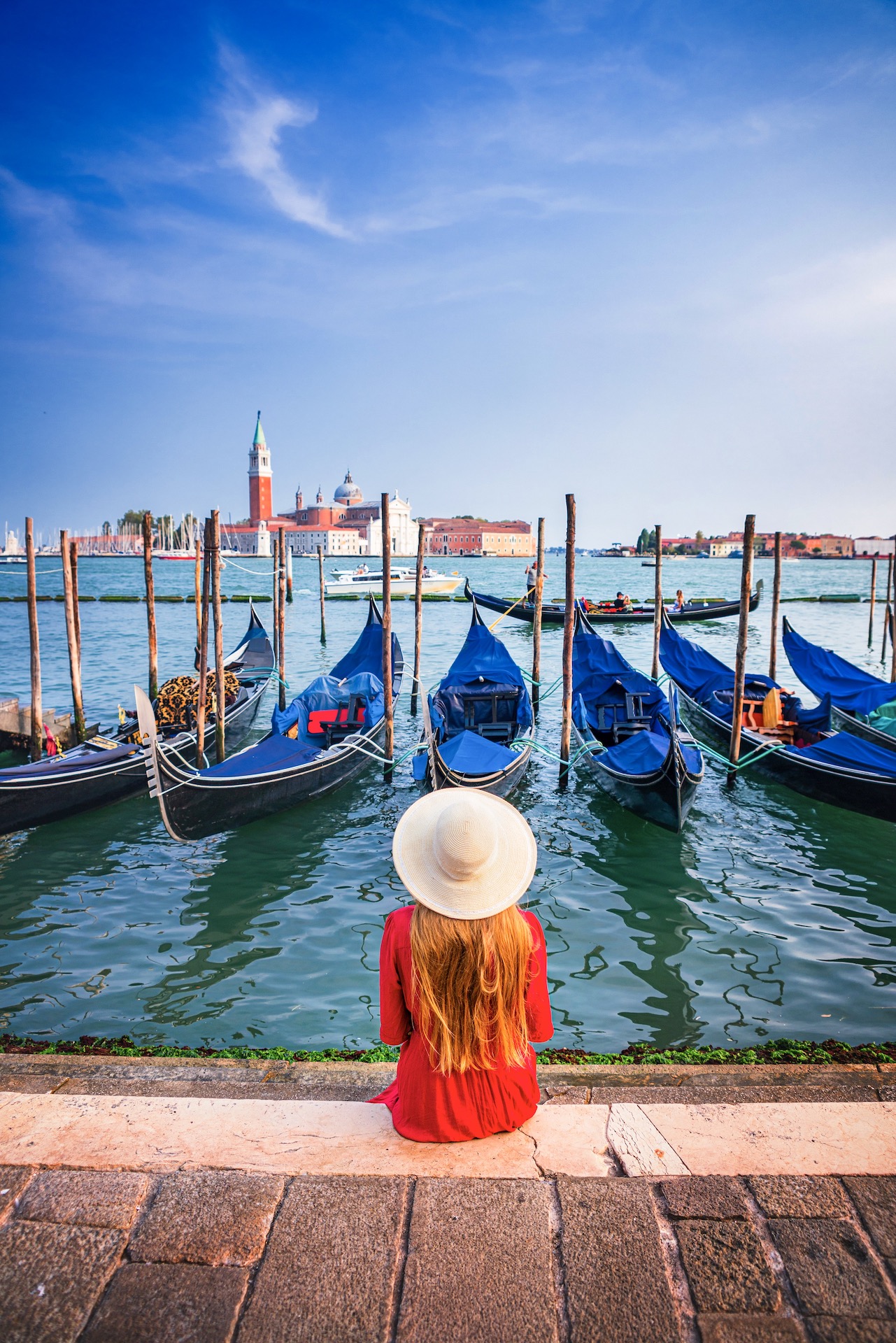 woman in red dress sitting along Venetian canal lined with gondolas on a Northern Italy itinerary.