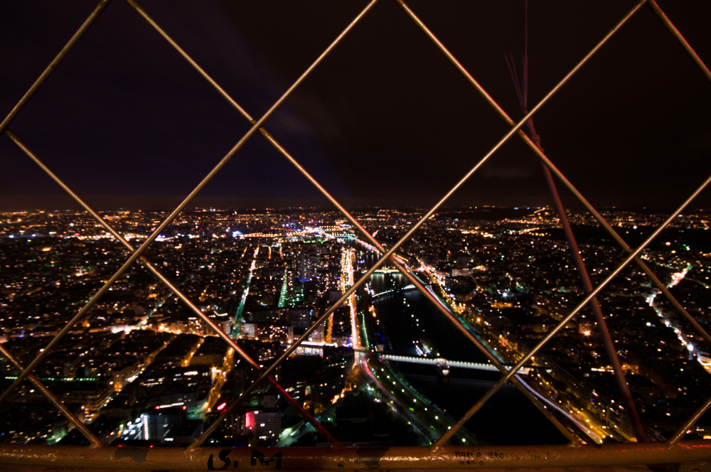 View from Eiffel Tower through fencing to the city lights below.
