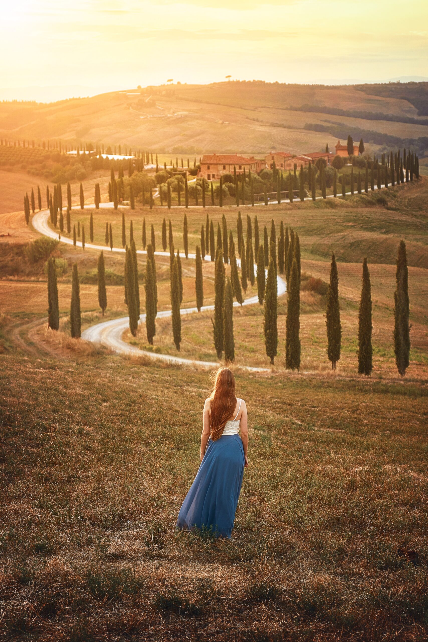 Woman in a blue skirt stands looking out at a winding Tuscany road and distant villa at golden hour.