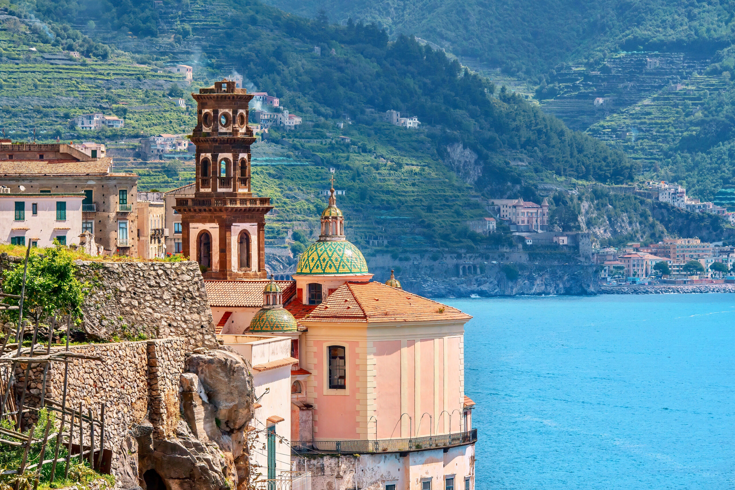 Photo of Castiglione di Ravello beach. A bell tower is shown next to an exquisite green and yellow majolica tiled dome church. Light blue water and green mountains are seen in the background. 