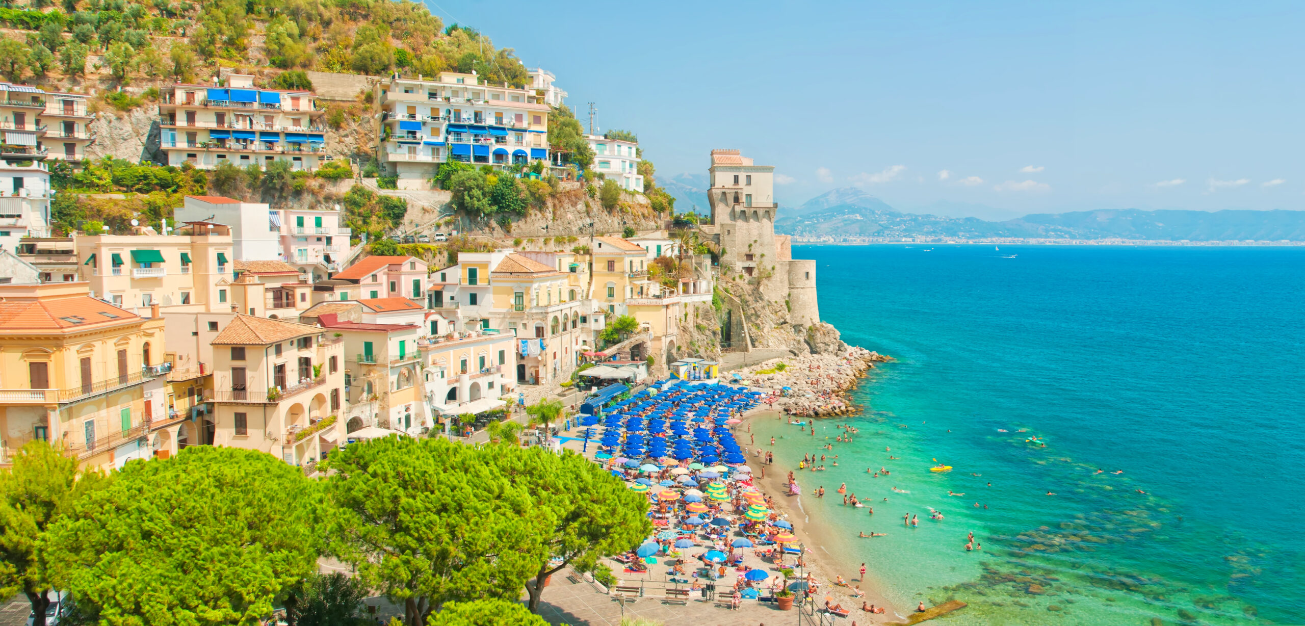 Photo of Cetara Beach. Lush green landscaping bordering a white sand beach. Several blue umbrellas are shown with a stone tower in the background. 