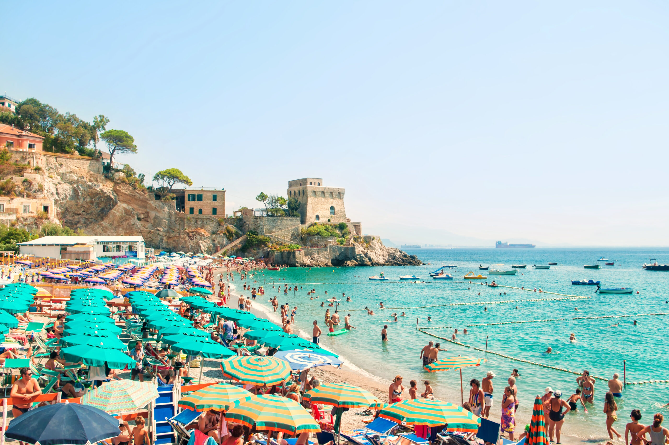 Photo of Erchie beach showing people sunbathing in aqua water with a stone tower in the background.