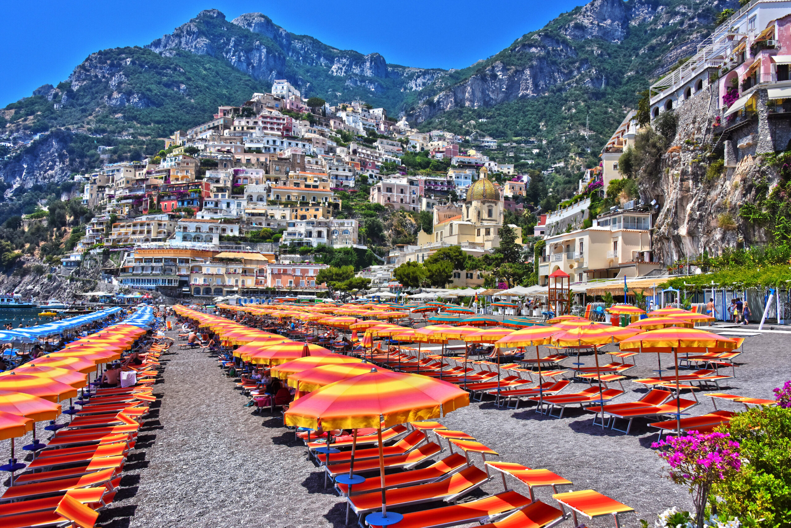 Photo of Marina Grande of Positano on the Amalfi Coast. Orange, yellow, and pink umbrellas are in the forefront with rocky cliffs in the background.