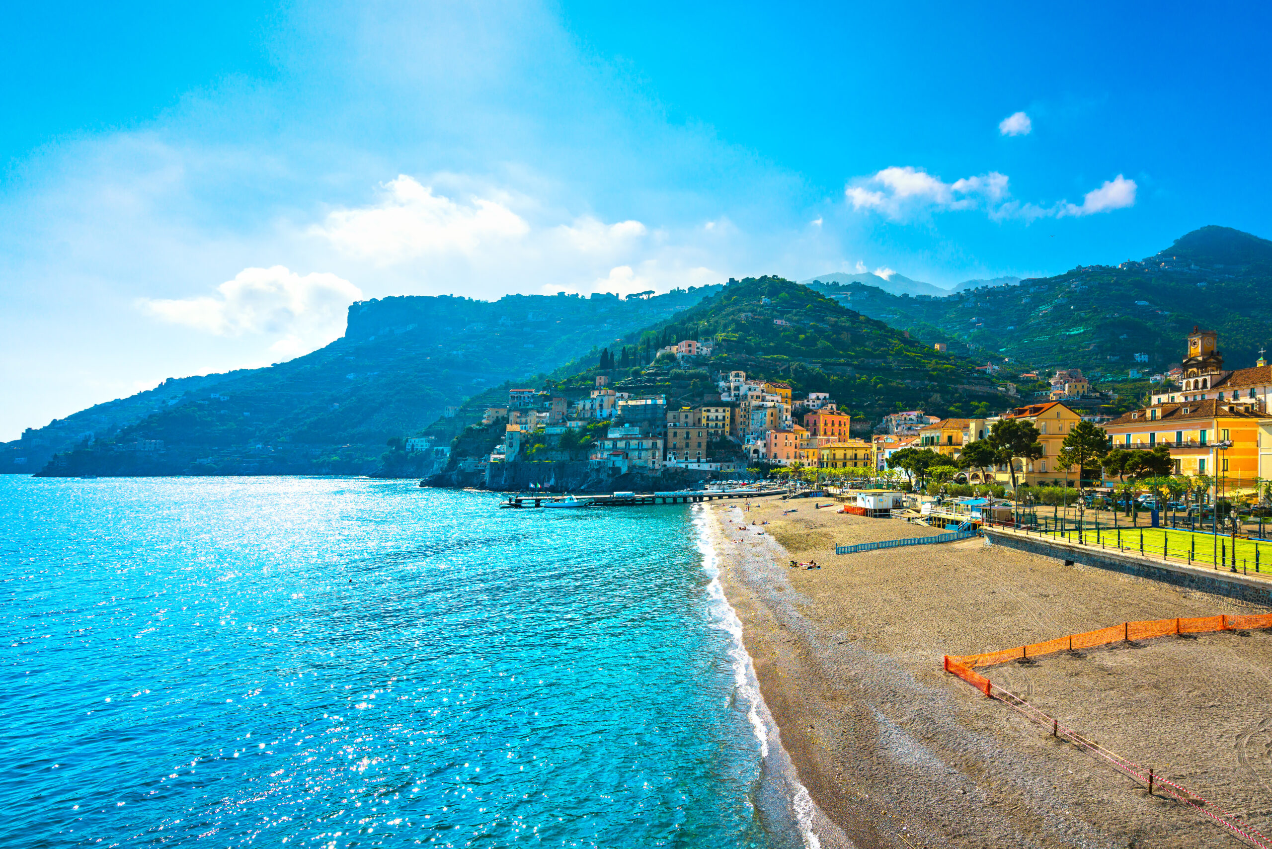 Photo of Minori beach. Crystal blue water is seen meeting a sandy beach. Colorful resort buildings line the beach. A prominent green covered mountain is in the main background.