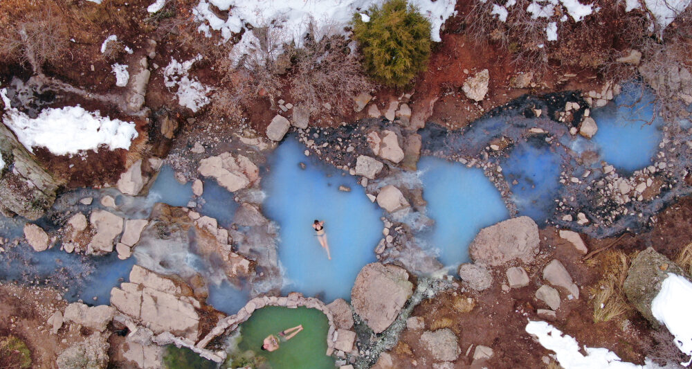 A sky view of two bathers in a milky blue hot springs surrounded by snow covered rocks.