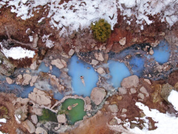 A sky view of two bathers in a milky blue hot springs surrounded by snow covered rocks.