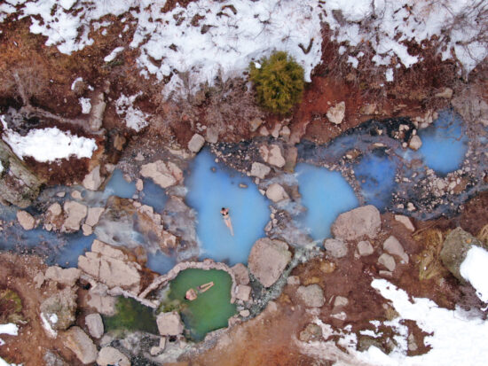 A sky view of two bathers in a milky blue hot springs surrounded by snow covered rocks.