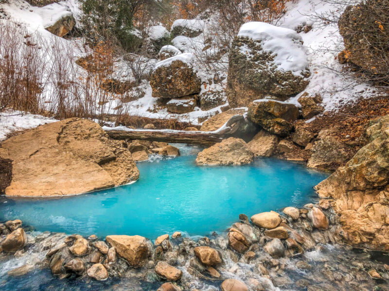 Diamond Fork Hot Springs in Utah. An aqua colored hot spring sits surrounded by snow covered rocks. 