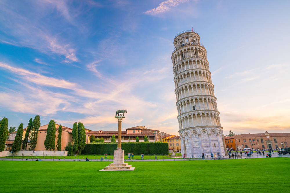 The Leaning tower of Pisa against a brilliant blue sky. You can see lots of buildings and people walking around. 