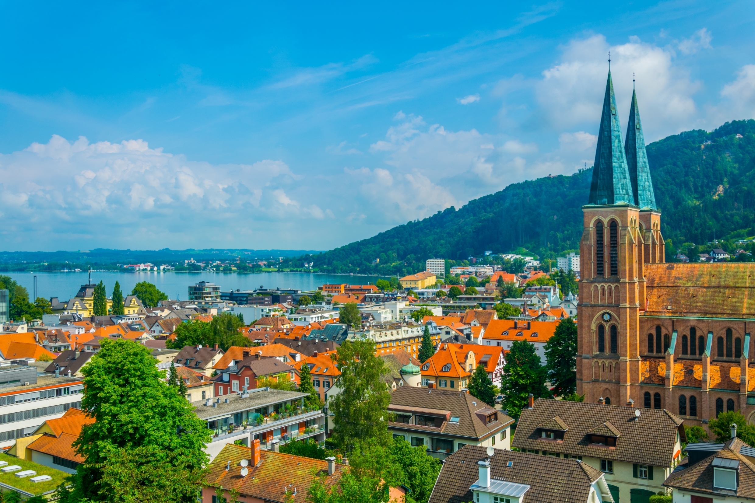 Photo of the small Austrian town Bregenz. The very unique double steeple church is shown on the right with many buildings and homes scattered on the left. Light blue water is far in the background with a green mountain on the right. 