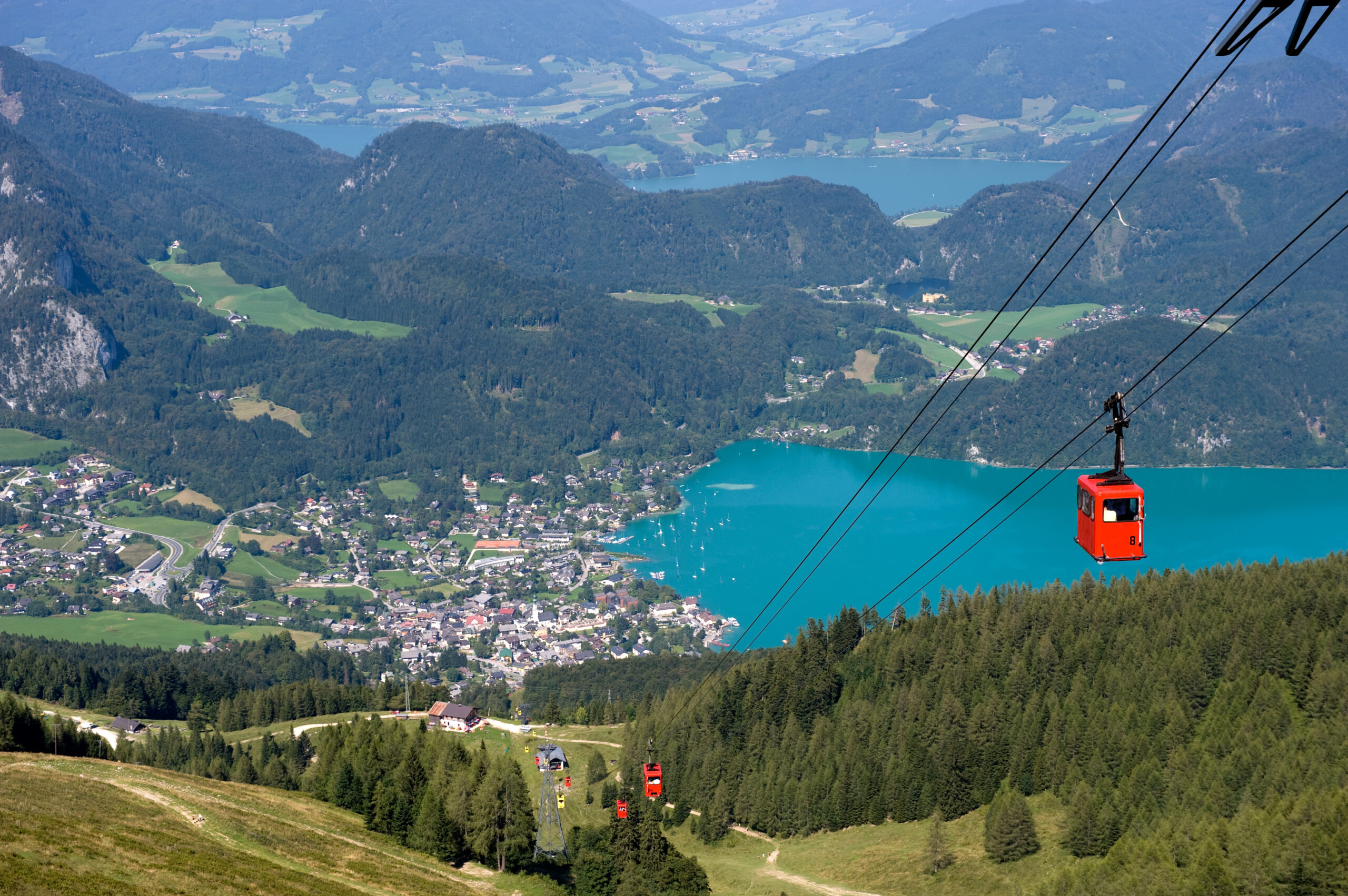 Photo of bright red cable car ascending an Alpine mountain. Small Austrian town with mountains and a brilliant blue lake are featured in the background.