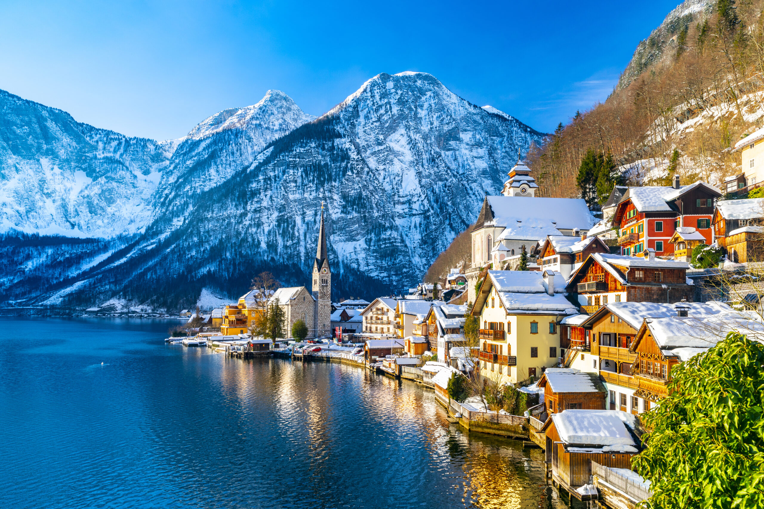 Photo of Hallstat, a small town in Austria, during winter. Colorful snow covered cottages are situated alongside a beautiful lake with a massive snowy mountain in the background.