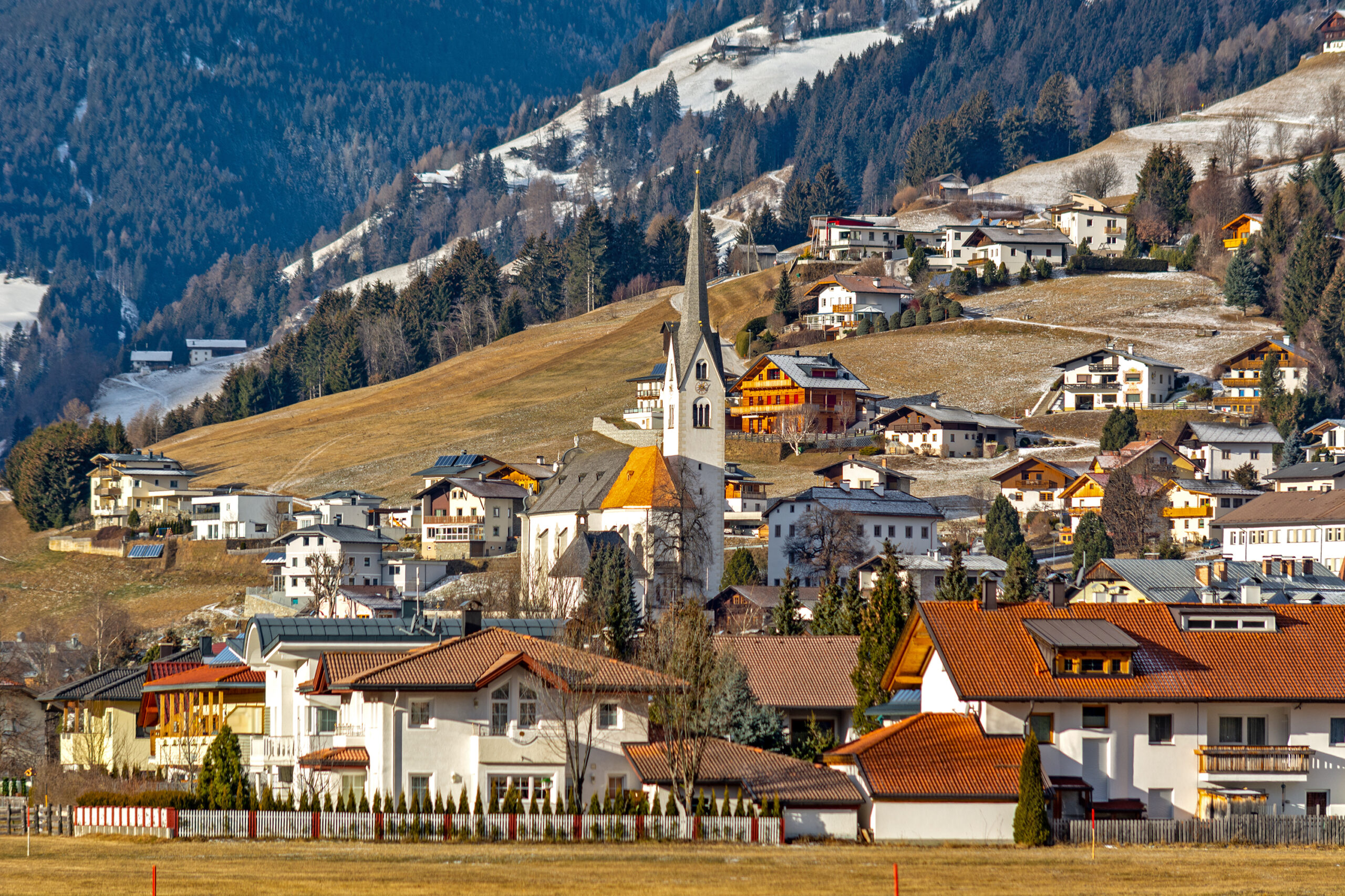 Photo of the small Austrian town Lienz. Homes are scattered at the base of a mountain. A church is seen right in the center of the town. Snowy mountains are in the far background as the elevation increases. 