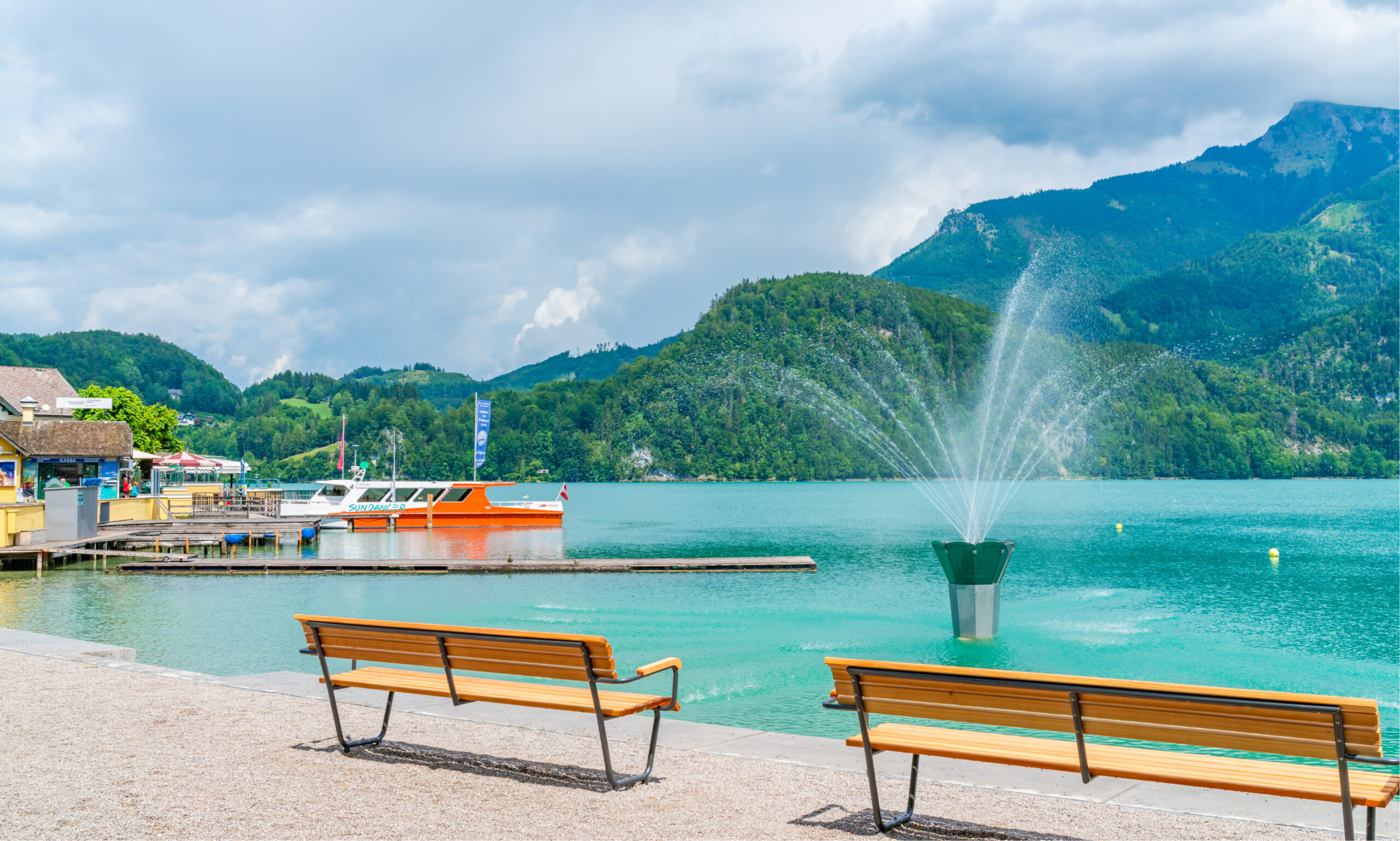 Photo of the stunning blue waters of the Wolfgangsee in St. Gilgen. A small shop with a dock and boat are featured on the left. Two wooden benches are in the foreground facing the Wolfgangsee water that has a pretty fountain in the middle. Lush green mountains are in the background.