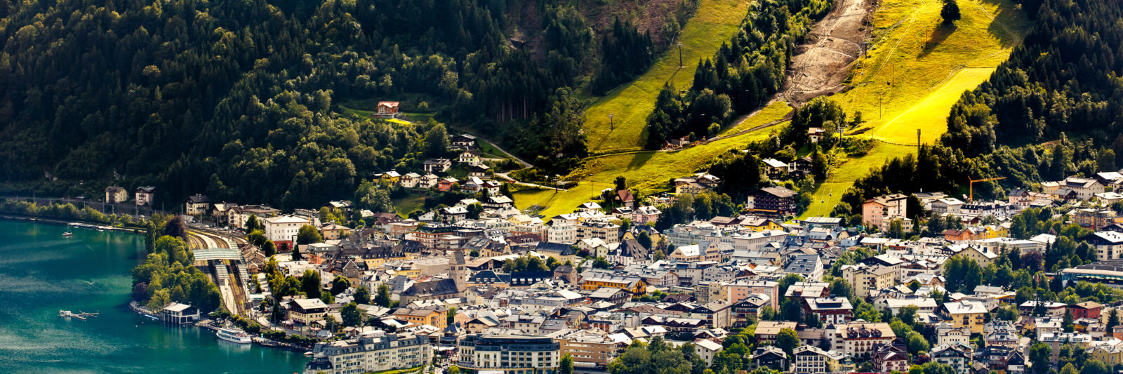 Photo featuring an amazing aerial view of Zell am See. Blue-green water is in the foreground. There are many waterfront resorts seen with just as many homes and buildings behind them. Light green grass is shown on large ski runs going up the mountain side. Dark green trees are on either side.