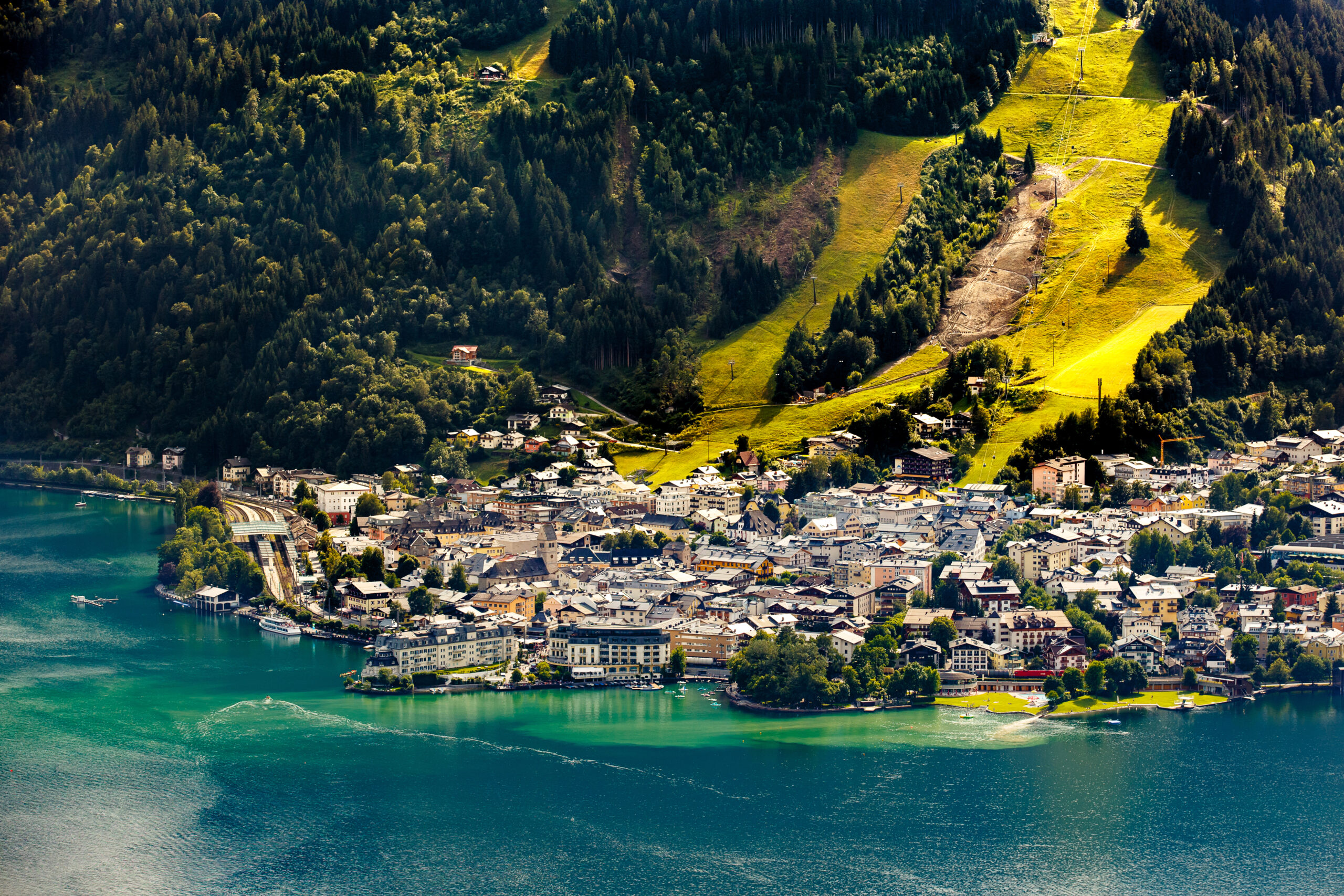 Photo featuring an amazing aerial view of Zell am See. Blue-green water is in the foreground. There are many waterfront resorts seen with just as many homes and buildings behind them. A few boats are tied off on docks. Light green grass is shown on large ski runs going up the mountain side. Dark green trees are on either side. 