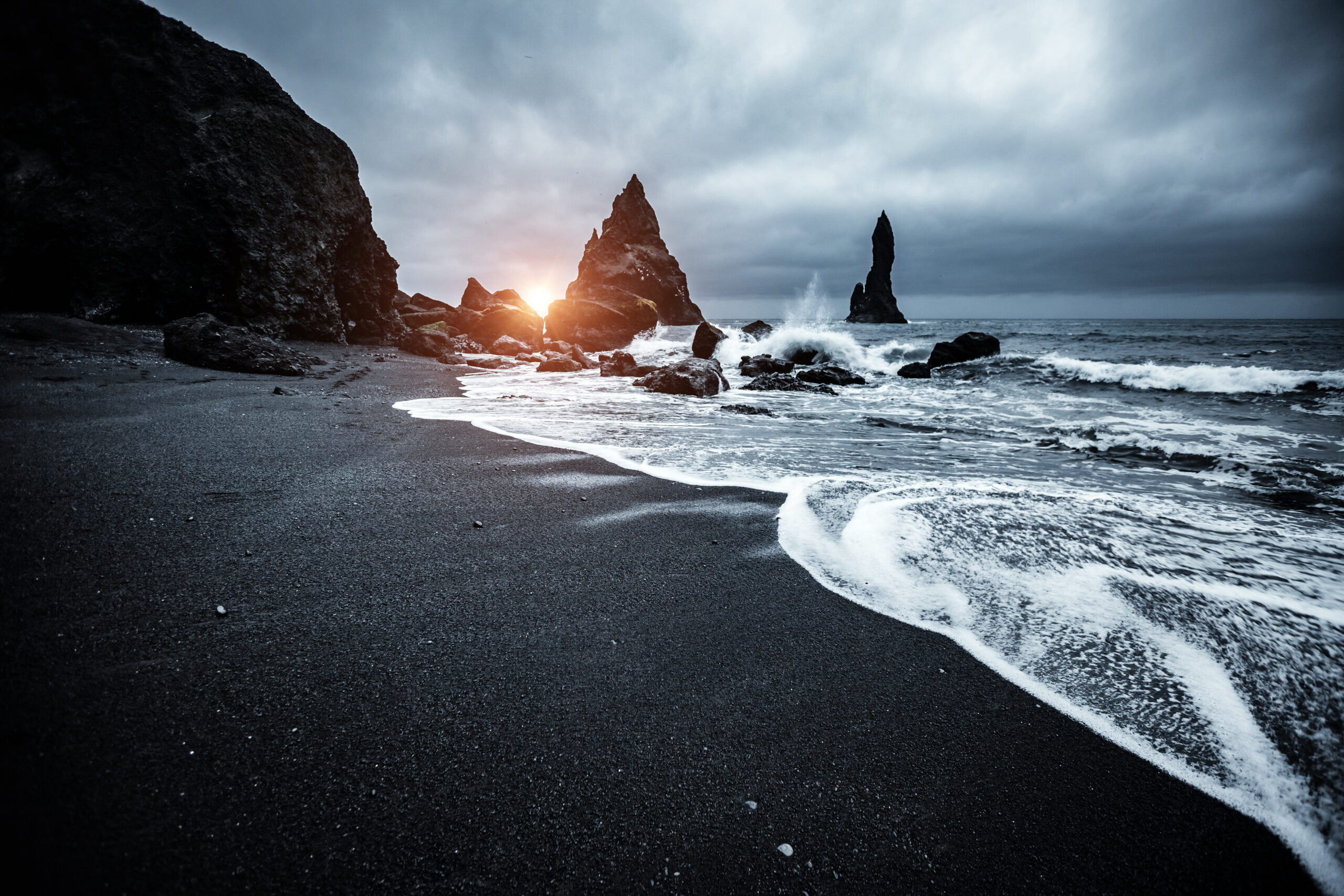 Photo of Reynisfjara Beach, Iceland. A black sand beach is seen with foamy tide washing inland. Rock formations are seen in the background.