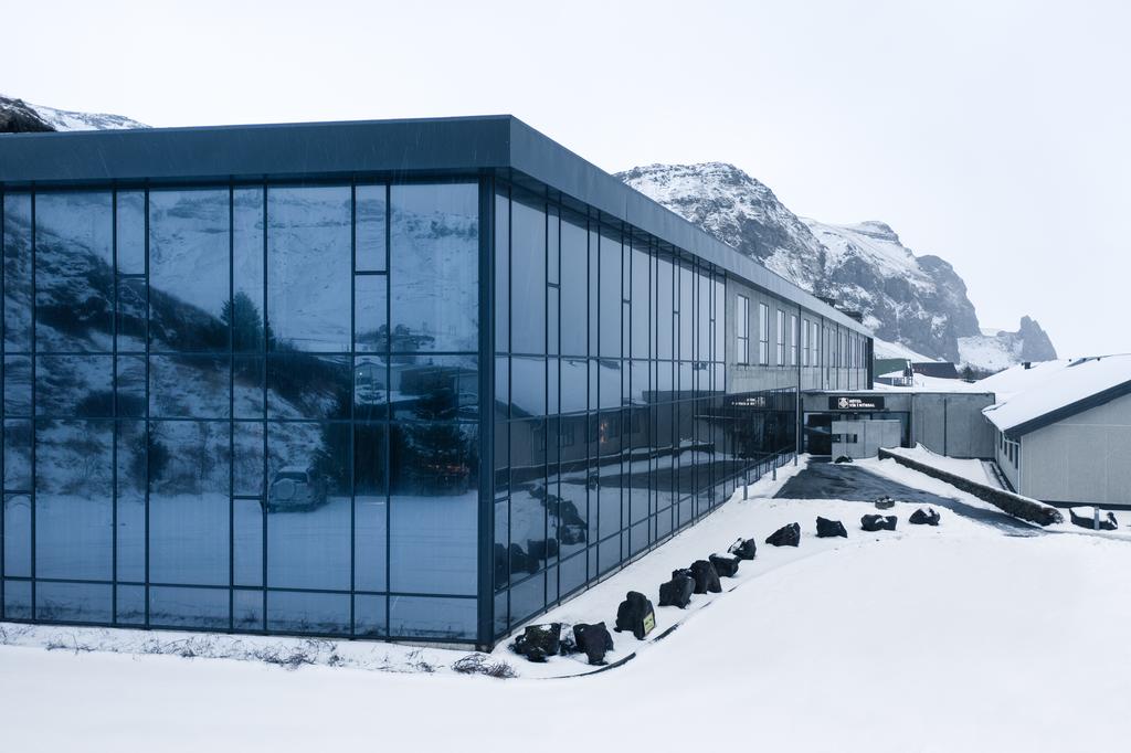 Photo of Hotel Vík í Mýrdal with gray blue glass windowed walls set against a snowy mountain backdrop and foreground. 