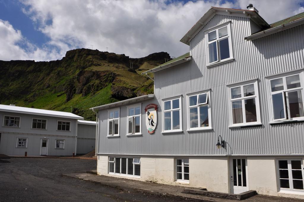 Photo of Puffin Hostel in Vik Iceland. A gray and white building is seen with lush green mountains in the background.