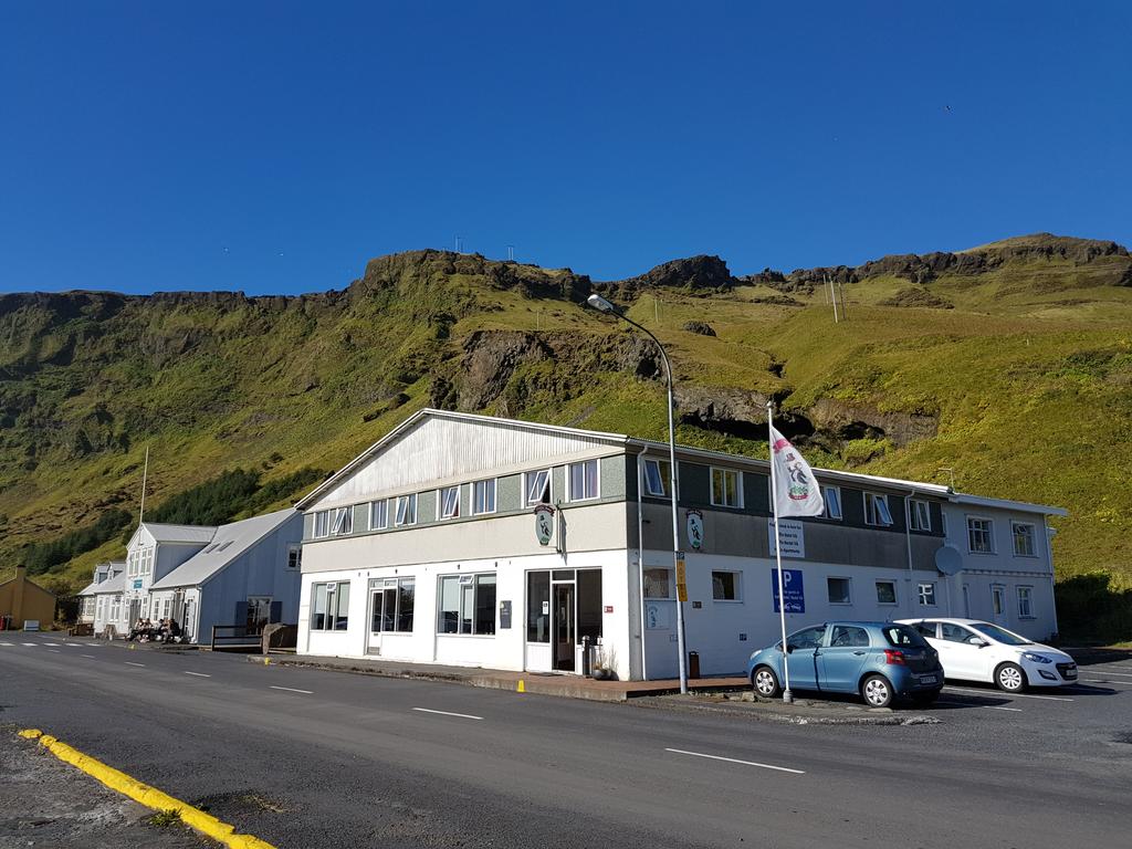 Photo of Puffin Hotel located in Vik Iceland. Small gray and white building is seen with a small mountain in the background. 