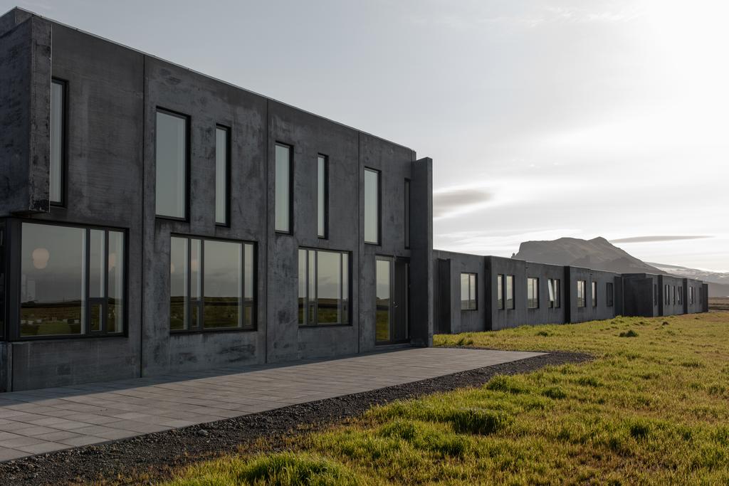 Photo of the exterior of Umi Hotel located in Vik Iceland. The building is dark gray and very rectangular. Mountains are seen in the distance. 